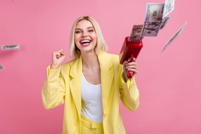 Femme souriante en tailleur jaune lançant des billets avec un pistolet à argent sur fond rose, illustrant la valorisation d’entreprise et la réussite entrepreneuriale